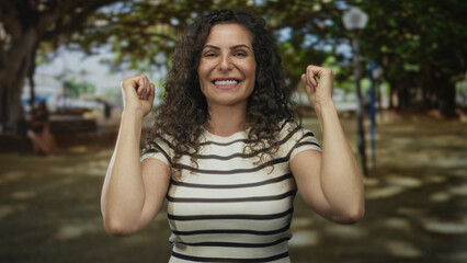 Smiling young hispanic woman raises clenched fists in sunlit bright green park at midday;...