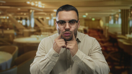 Man pinches cheeks in indoor restaurant dining area while wearing glasses and beige shirt;...