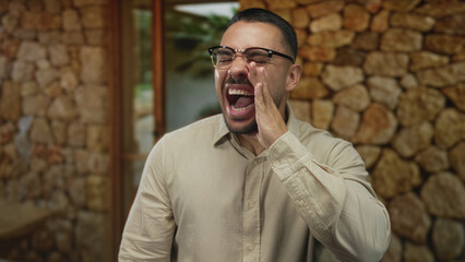 Hispanic man with hand to mouth shouting at building entrance; excitement urgency communication expression.
