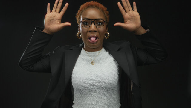 Young woman in white top and black jacket sticks tongue out with raised hands against black backdrop in studio; playful mischief.