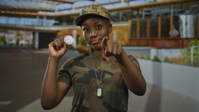 Woman in camouflage uniform holds a vote badge and points finger to badge on city street; civic duty.