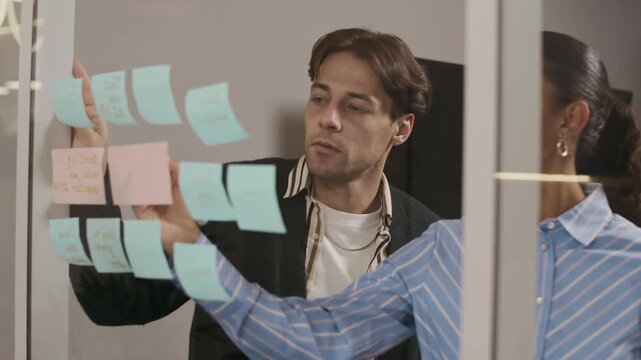 Male and female coworkers looking at multicolored stickers on polished window and discussing workday tasks