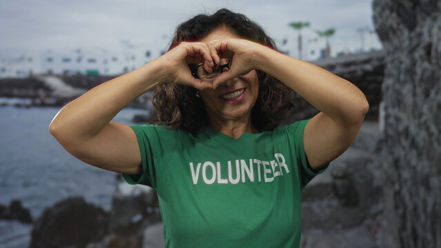 Woman smiling forms heart with hands by seaside wearing volunteer shirt on beach revealing supportive community spirit outdoors in nature. - Powered by Adobe