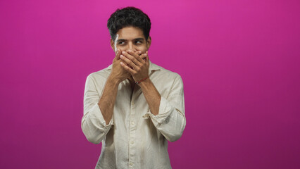 Obraz premium Young hispanic man hands covering mouth in studio with pink backdrop wearing beige linen shirt and small bracelet; playful surprise.