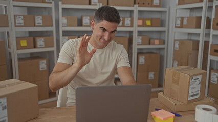 Man waving hand in building at laptop and parcels, gesturing hello to camera; friendly small business.
