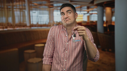 Man holding keys with outstretched hand, smirking and leaning by curved booth seating inside a lit building with wooden floor and pendant lamps; doubt.