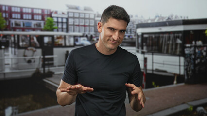 Fototapeta premium Man gesturing hands out palms down on street by canal boat in amsterdam; confidence casual friendliness.