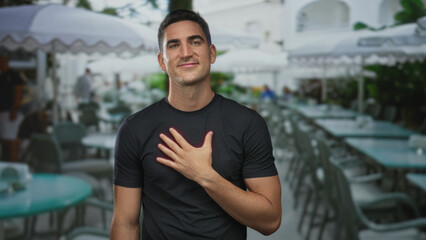 Man with hand on chest at outdoor restaurant terrace on the street, smiling to camera with calm expression; sincere gratitude.