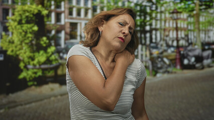 Woman holds shoulder with pained expression on city street lined with green trees and parked scooters; pain.