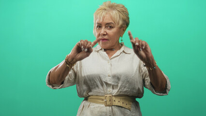 Senior hispanic woman wearing white blouse and beige belt points index fingers upward in studio with green backdrop, hands forward; firm warning.