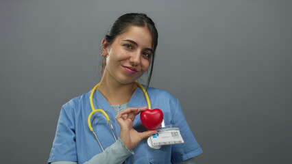 Young hispanic woman doctor holding heart model smiling confidently in medical uniform with stethoscope against isolated grey background suggesting health care and compassion.