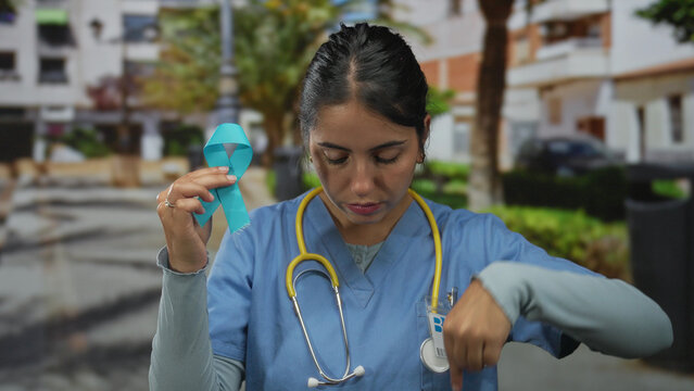 Young woman doctor wearing stethoscope holds blue ribbon and points finger down on street; compassionate support.