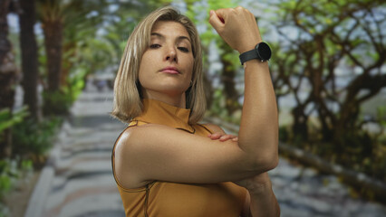 Bright blonde woman flexes bicep in park walkway under sunlight filtering through leaves; empowerment.
