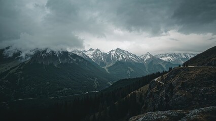 Fototapeta premium Snow-capped mountains under stormy clouds with dense forests and rugged terrain. Mountain landscape, wilderness, scenic nature view.