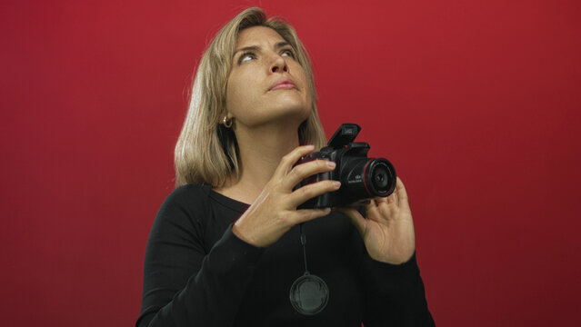 Woman removes lens cap and adjusts digital camera lens and squints eye in vivid red studio; creative focus.
