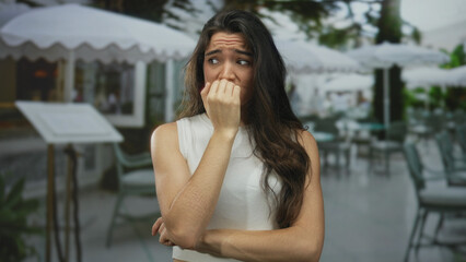 Young hispanic woman biting fingernails with worried gaze on restaurant terrace; anxiety worry uncertainty.