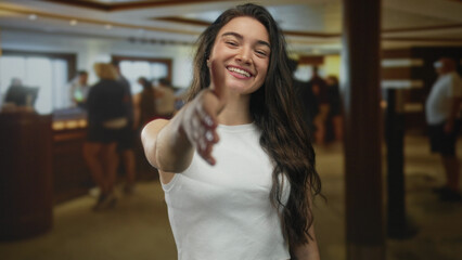 Young hispanic woman wearing white tank top extends bare hand for handshake in hotel building reception; friendliness.