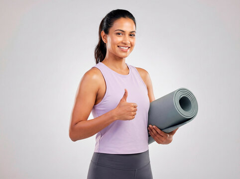 A happy, fit young indian woman in sportswear stands holding a yoga mat and giving a thumbs up gesture, ready for her workout or exercise class