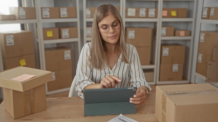 Woman inspects small cardboard package with hands at desk using tablet in office building;...