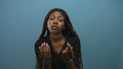Woman with braided hair wearing a cross necklace showing open hands and palms up gesture in a studio against a blue wall; pleading apology.