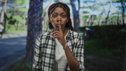 Fototapeta premium African american woman with braided hair smiling, hand raised pointing toward camera in park forest setting, wearing plaid shirt and white tee; confidence empowerment.