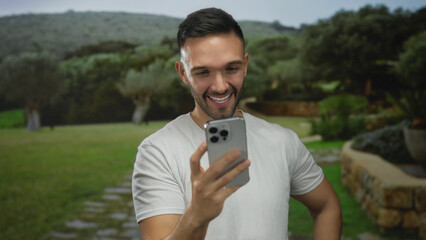 Young man smiling at smartphone in lush green park, enjoying outdoors with trees and stone path.