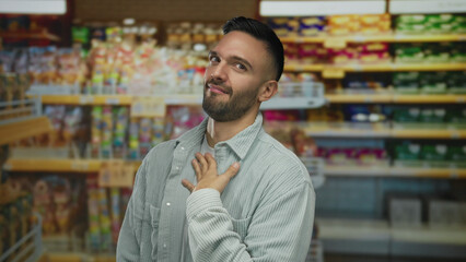 Young hispanic man in grocery store expressing emotions surrounded by various colorful products on shelves creating a vibrant shopping environment backdrop