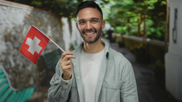 Young hispanic man smiling and holding a swiss flag outdoors in a green park setting, showcasing his casual attire and cheerful expression.