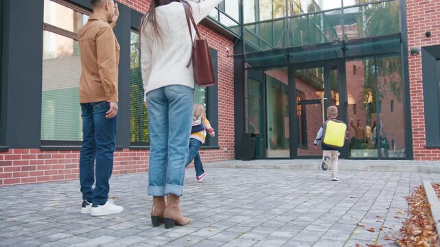 Parents standing on paved path waving as two kids with backpacks run toward school entrance. Family supporting children beginning day at bright brick campus. Morning routine. School drop off.