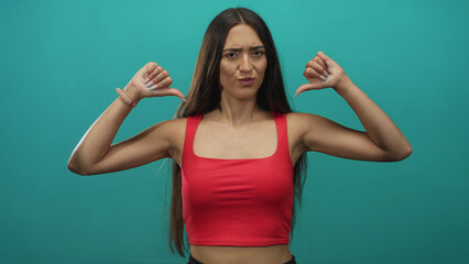 Young hispanic woman pointing both thumbs to chest in teal studio wearing red crop top and smartwatch, arms raised; skepticism attitude.