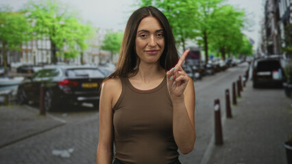 Woman with finger to eye on a city street wearing brown tanktop and long hair, slight smirk and raised hand gesture; playful confidence.