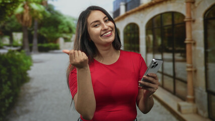 Woman in red shirt holding smartphone, beckoning with hand near a building entrance on a street while smiling at camera; happy friendly invitation.