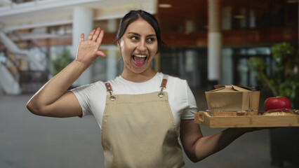 Woman waitress holding wooden tray with takeaway box and apple, waving hand in building; friendly service hospitality.
