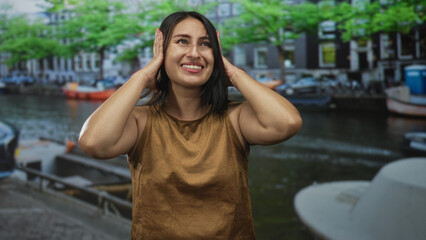 Fototapeta premium Woman holding hands to ears on a street by an amsterdam canal, smiling with closed eyes; playful joy.
