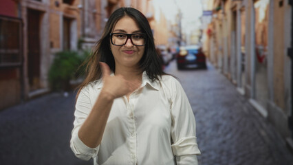 Obraz premium Woman on street in white shirt and glasses with hand to chest and dismissive smirk, standing in narrow city lane; annoyance confidence.