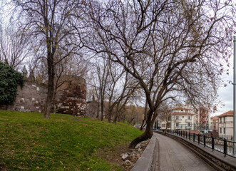 A pedestrian alley at the base of Hisar Park hill, following the ancient fortress wall with round defensive towers. Bare trees on green lawn above residential streets. Ankara, Turkey.

