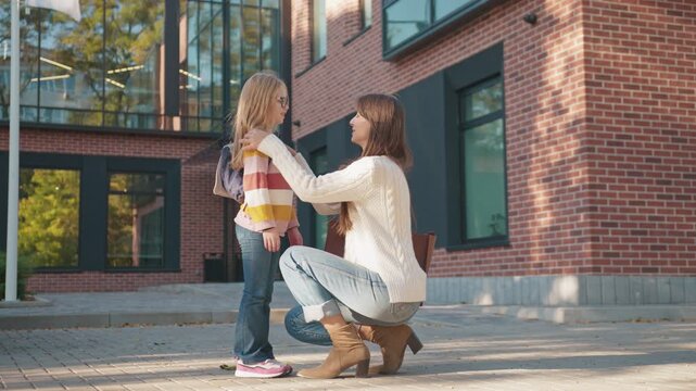 Female parent kneeling adjusting schoolgirl backpack while talking on brick walkway. Woman checking straps supporting child before leaving campus. Girl standing calmly during quick preparation.