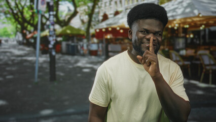Fototapeta premium Young man smiling points finger and finger to lips for silence while standing near outdoor cafe on a city street; playful secrecy.