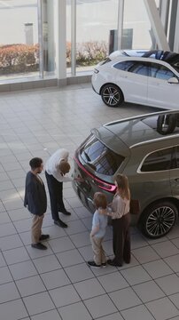 Vertical wide angle shot of family choosing new SUV car in dealership showroom with car salesman assisting and opening car trunk