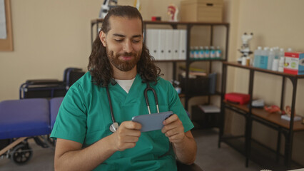Man doctor with stethoscope holding smartphone and tapping screen in clinic room, seated by treatment couch and shelving of supplies; calm concentration.