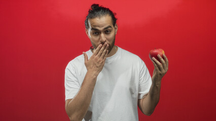 Man holding red apple with hand to mouth in red studio, smiling and offering fruit; playful temptation.