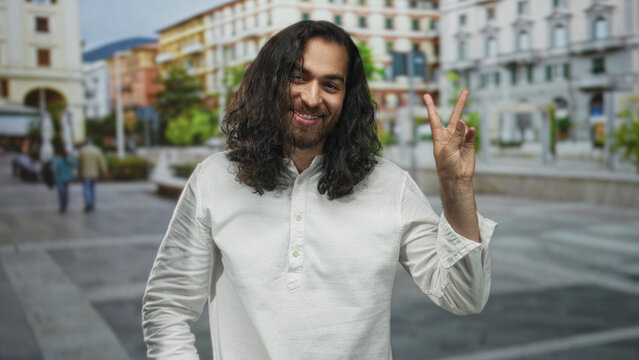 Young arab man flashing peace sign with bare hand in sunny city street plaza among buildings and passersby; peaceful optimism. - Powered by Adobe
