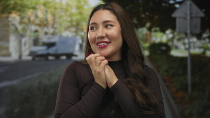Woman with long dark hair clasps hands with gentle smile on tree lined city street at golden hour; gratitude.