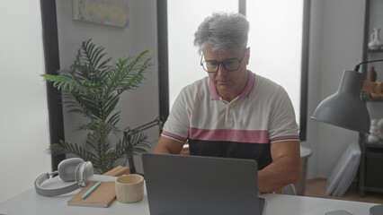 Man adjusting glasses while typing on a laptop at a desk in a studio with lamp and plant; quiet concentration.