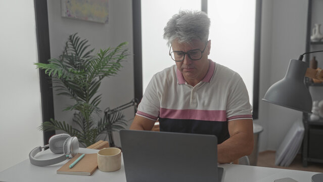 Man adjusting glasses while typing on a laptop at a desk in a studio with lamp and plant; quiet concentration.