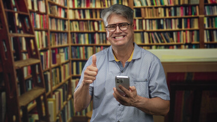 Senior man with grey hair and glasses taps a smartphone with thumb while smiling amid tall library shelves in a building aisle; contentment connection.