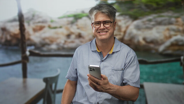 Man holding smartphone and tapping screen on a seaside terrace building, wearing glasses and short sleeve shirt while smiling and looking aside; relaxation leisure connection.