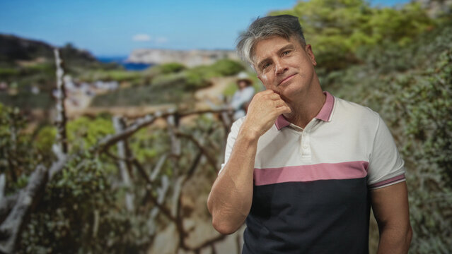 Man touching cheek with hand, head tilted near a wooden fence in a studio coastal set with shrubs and distant sea; quiet contemplation.