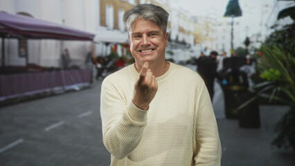 Senior man smiling, beckoning with finger on a street market beside stalls and planters; playful invitation.