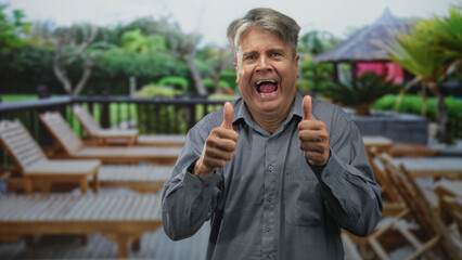 Man cups hand to ear and laughs beside lounge chairs on a resort building deck, visible ear and hand gesture; playful curiosity.